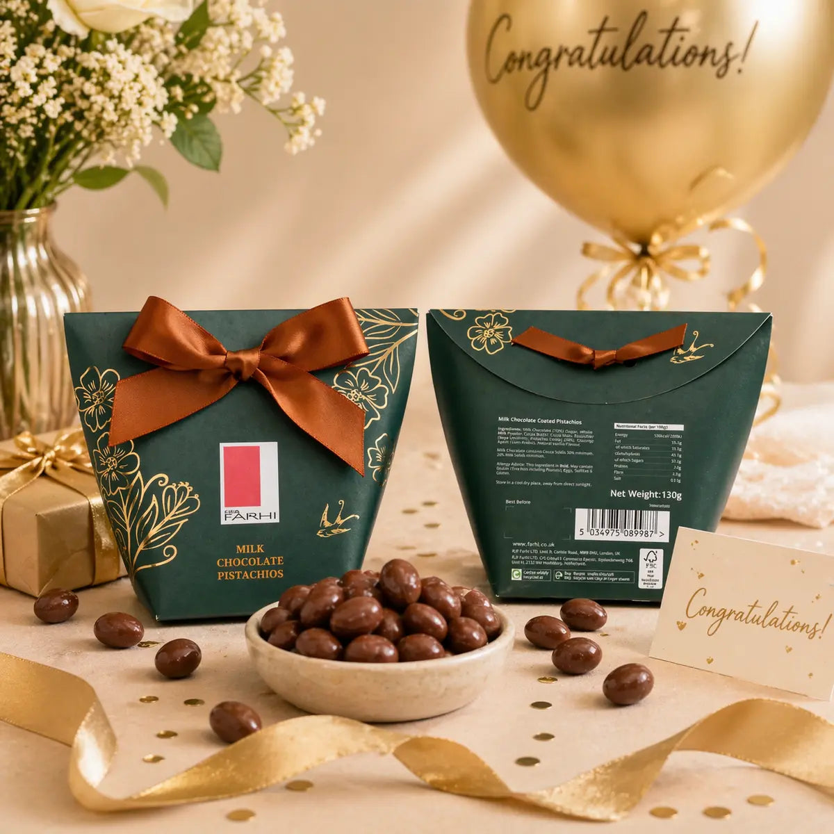 Two green chocolate boxes with brown ribbons on a table with flowers, balloons, and a card.